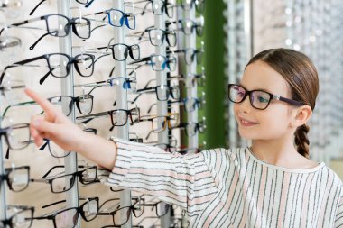 positive child pointing at assortment of eyeglasses in optics shop