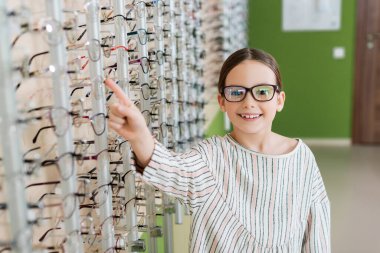 happy girl pointing with finger while choosing eyeglasses in optics shop