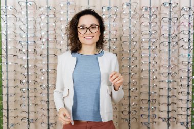cheerful woman looking at camera near assortment of eyeglasses in optics shop