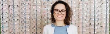 pleased young woman smiling at camera near assortment of eyeglasses in optics shop, banner