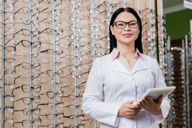 happy asian oculist with digital tablet looking at camera in optics shop