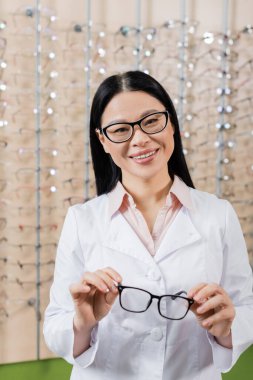 cheerful asian ophthalmologist looking at camera while holding eyeglasses in optics store