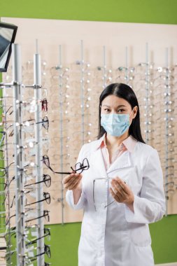 asian oculist in medical mask comparing eyeglasses while working in optics shop clipart
