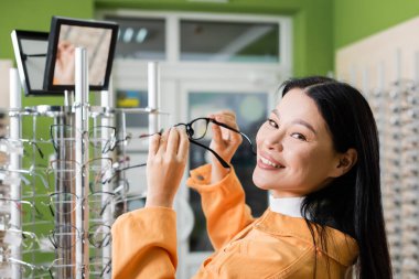 asian woman smiling at camera while holding eyeglasses near mirror in optics shop