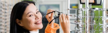 happy asian woman looking at camera while choosing eyeglasses in optics salon, banner