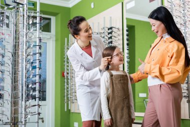 asian woman showing thumb up near happy daughter in eyeglasses and smiling oculist in optics store