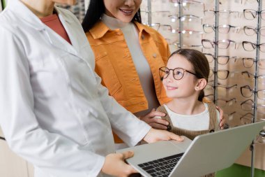pleased child in eyeglasses smiling near mother and oculist with laptop in optics shop