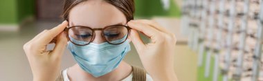 oculist putting eyeglasses on girl in medical mask, banner