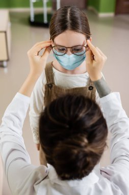 blurred ophthalmologist trying eyeglasses on child in medical mask in optics store