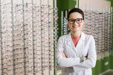 pleased ophthalmologist in white coat and eyeglasses standing with crossed arms in optics store
