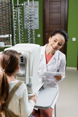 smiling oculist with digital tablet testing eyesight of child on vision screener in optics store