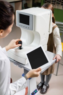 blurred ophthalmologist holding digital tablet with blank screen while measuring eyesight of girl on autorefractor in optics store