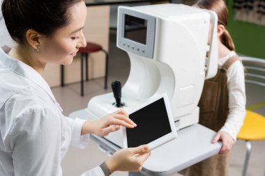 smiling oculist using digital tablet with blank screen while measuring vision of blurred girl on ophthalmoscope