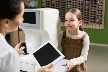 blurred oculist holding digital tablet with blank screen near positive girl and vision screener in optics store
