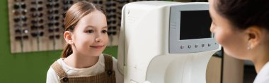 smiling girl looking at blurred oculist near vision screener in optics salon, banner clipart