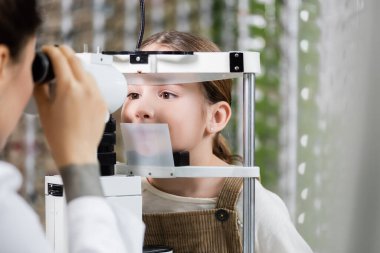 blurred optometrist measuring eyesight of girl on vision screener in optics shop clipart