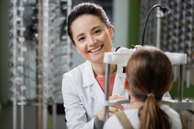 pleased oculist looking at camera while checking eyesight of girl on vision screener in optics shop