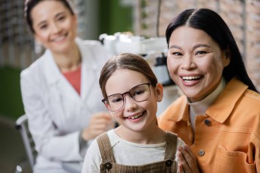happy kid in eyeglasses looking at camera near asian mom and blurred ophthalmologist in optics shop