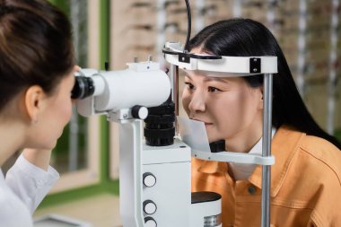 blurred optometrist testing eyesight of asian woman on vision screener in optics shop clipart