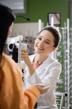 young optometrist smiling near blurred woman and ophthalmoscope in optics shop
