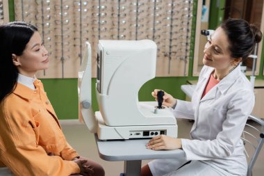 smiling optometrist looking at asian woman near vision screener and blurred assortment of eyeglasses