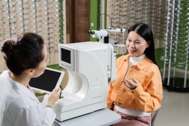 smiling asian woman pointing with hand near blurred oculist with digital tablet and vision screener in optics shop