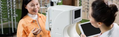 smiling asian woman pointing with hand near vision screener and oculist with digital tablet, banner
