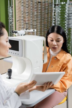 blurred oculist showing digital tablet to asian woman near vision screener in optics salon