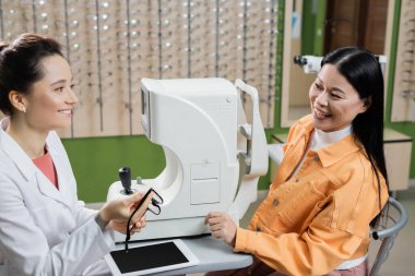 smiling optometrist holding eyeglasses near happy asian woman and ophthalmoscope in optics shop