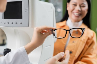 selective focus of eyeglasses in hand on oculist near vision screener and smiling asian woman