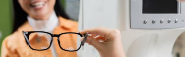 partial view of oculist holding eyeglasses near smiling woman and blurred vision screener, banner