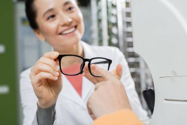 woman pointing with finger at eyeglasses in hand of happy oculist in optics shop