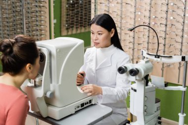 asian optometrist checking vision of blurred woman on ophthalmoscope in optics store
