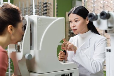 asian oculist pointing at vision screener while measuring eyesight of blurred woman in optics store