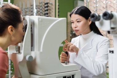 asian oculist pointing at vision screener while measuring eyesight of blurred woman in optics store clipart
