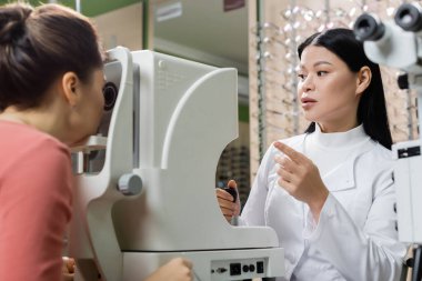 asian optometrist pointing with finger while measuring eyesight of blurred woman on vision screener
