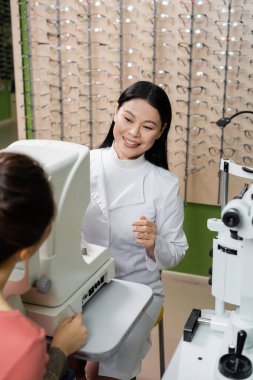smiling asian oculist testing vision of blurred woman on ophthalmoscope in optics shop