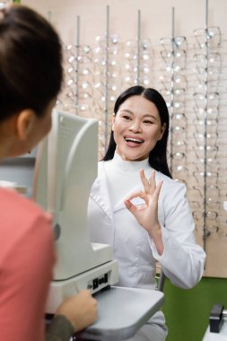 pleased asian oculist showing okay gesture near vision screener and blurred woman in optics salon