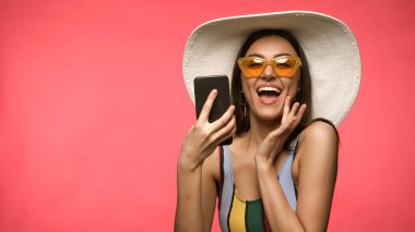 Güneş gözlüklü ve mayo giyen harika bir kadın. Pembe üzerine izole edilmiş cep telefonu kullanıyor. 