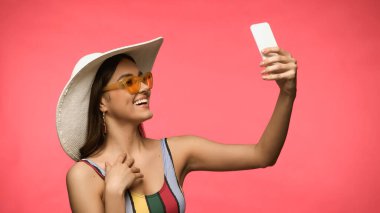 Mayo giymiş, güneş gözlüklü, gülümseyen bir kadın pembe bir telefondan selfie çekiyor. 