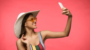 Güneş şapkalı mutlu kadın pembeden izole edilmiş akıllı telefondan selfie çekiyor. 
