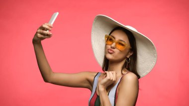 Güneş şapkalı ve güneş gözlüklü esmer kadın pembe renkte izole edilmiş akıllı telefondan selfie çekiyor. 