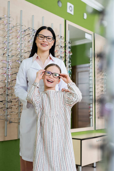 happy girl trying on eyeglasses near smiling asian oculist in optics shop on blurred foreground