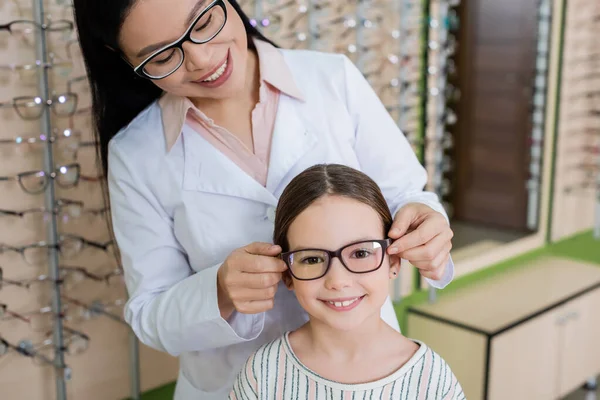 笑着的亚洲眼科医生为在光学商店看相机的快乐女孩选择眼镜 — 图库照片