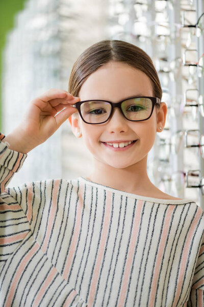 pleased child looking at camera while trying on eyeglasses in optics shop on blurred background