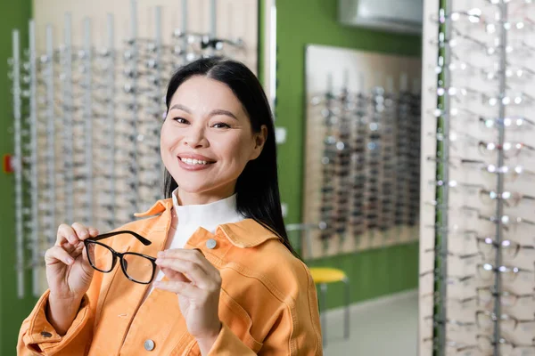在光学商店举着眼镜看着相机的亚洲女人高兴极了 — 图库照片