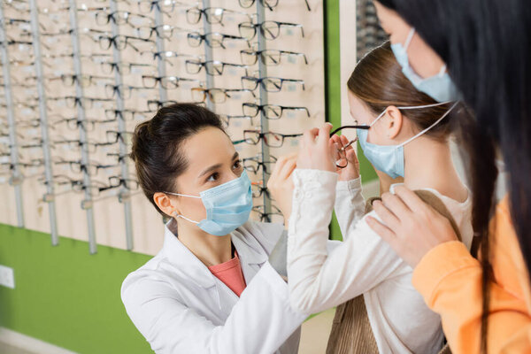 oculist in medical mask trying glasses on girl near blurred asian woman in optics store