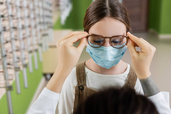 blurred ophthalmologist trying eyeglasses on girl in medical mask in optics salon