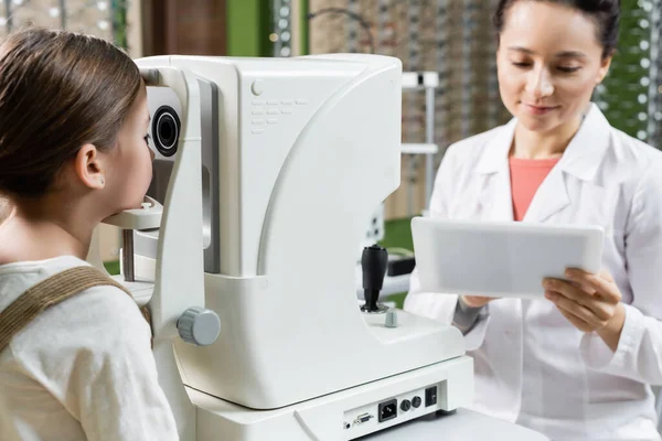 Positive Optometrist Looking Digital Tablet While Measuring Vision Girl Autorefractor — Zdjęcie stockowe