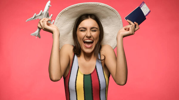 Cheerful tourist in sun hat and swimsuit holding passport and toy plane isolated on pink 